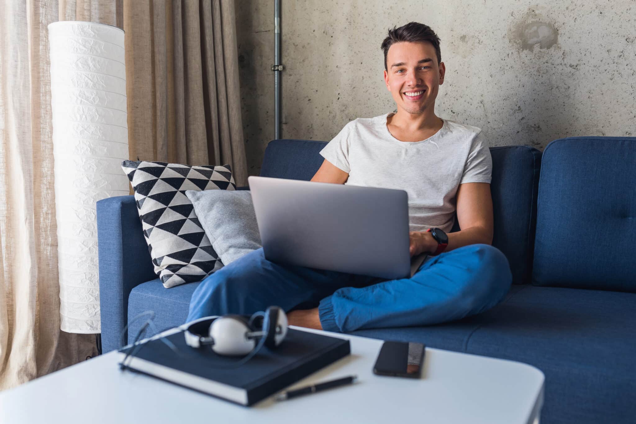 young attractive man sitting on sofa at home working on laptop online, using internet, smiling, happy mood, freelancer, free leisure time, relaxed, modern job lifestyle, looking in camera young attractive man sitting on sofa at home working on laptop online, using internet, smiling, happy mood, freelancer, free leisure time, relaxed, modern job lifestyle, looking in camera